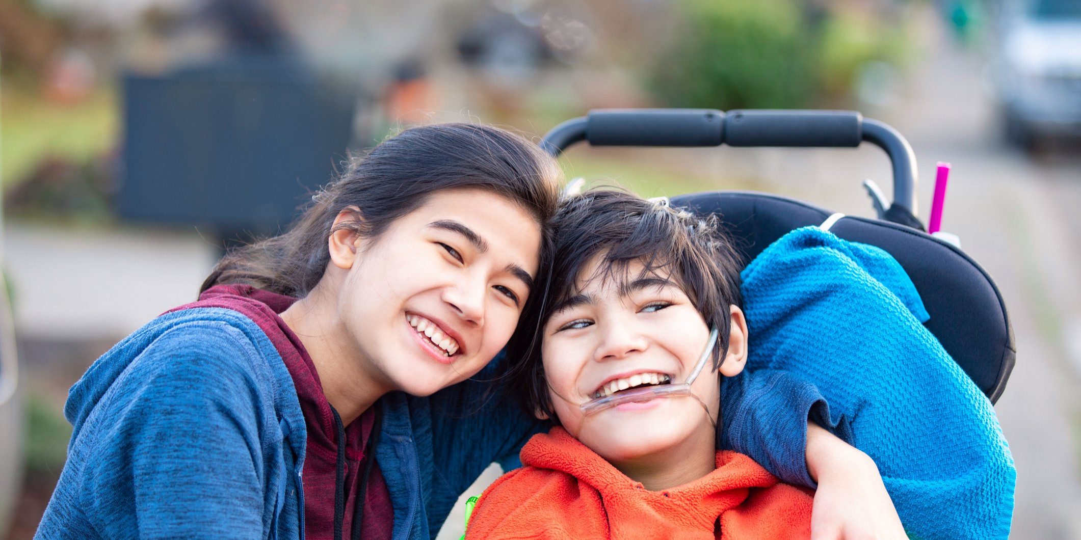 Biracial big sister lovingly hugging disabled little brother in wheelchairoutdoors, smiling